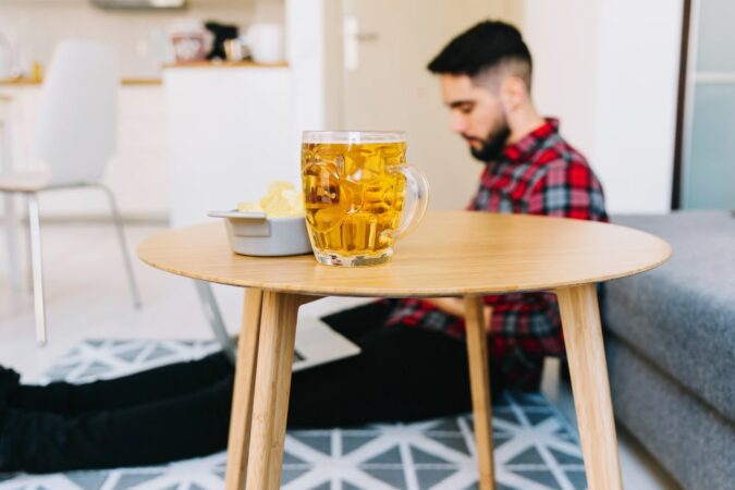 Chope de bière classique remplie de bière blonde sur une table basse en bois, avec un bol de chips et un homme en chemise à carreaux rouge en arrière-plan