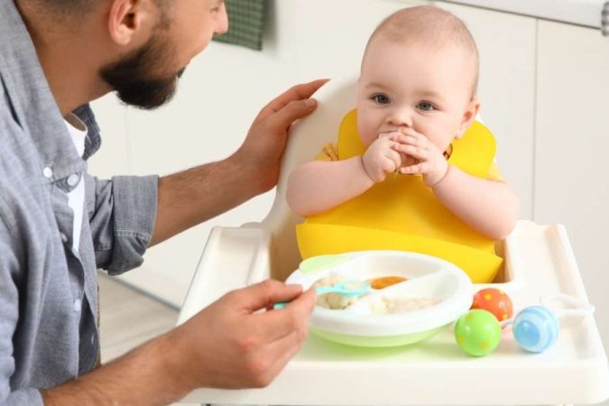 Un père nourrit son bébé dans une chaise haute, illustrant un mode de garde familial impliqué au quotidien.